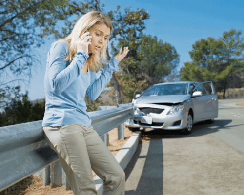 Woman looking stressed on the phone on the side of the road, behind her is her car crashed into the guardrail.