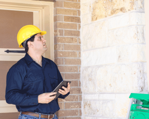 A man wearing a yellow hardhat and holding a clipboard does an inspection of a building.