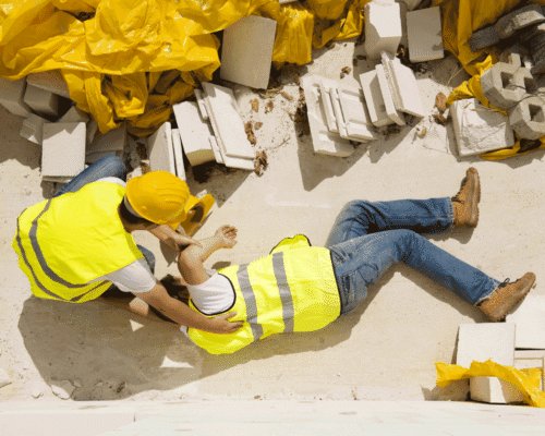 Two construction workers wearing safety vest and hard hats on a construction site. One is on the ground injured and the other is attending to him.