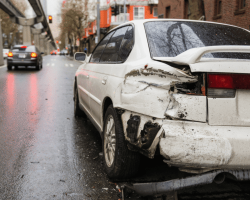 Rear view of a damaged white car parked on a wet city street, showing significant impact to the back corner and bumper. Other vehicles and a monorail track are visible in the background under overcast skies with red traffic lights reflecting on the pavement