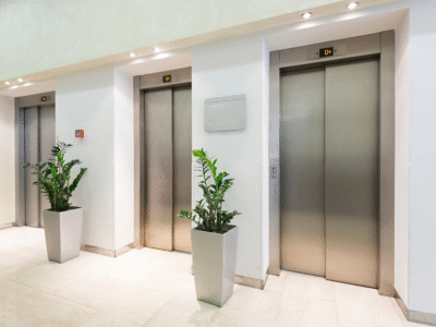 Modern indoor hallway with three stainless steel elevator doors, flanked by large potted green plants and illuminated by recessed ceiling lights. Each elevator displays the floor indicator reading '0+'
