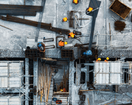 Construction workers in hard hats coordinating tasks on a busy building site viewed from above, surrounded by scaffolding, rebar, and tools