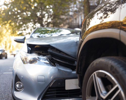 Close-up of a car accident showing the front end of a silver vehicle that has collided with the rear of a dark SUV on a city street. The silver car's hood is crumpled and its bumper is damaged, with other vehicles visible in the background