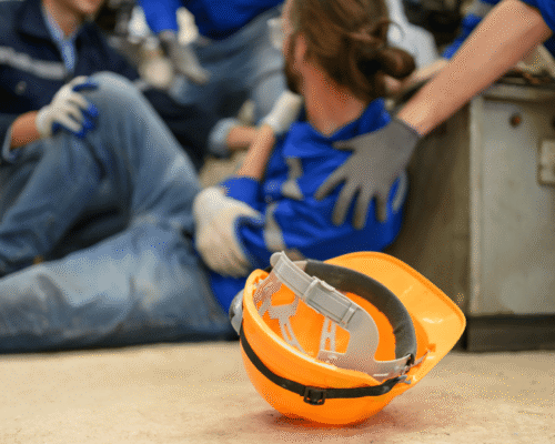 Close-up of an orange construction helmet on the ground with a blurred scene in the background showing a worker receiving assistance from colleagues after an apparent workplace injury. The injured worker is sitting on the floor wearing blue work clothes, surrounded by others wearing gloves