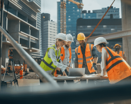 Construction crew in safety vests and hard hats reviewing plans at a job site with high-rise buildings and cranes in the background