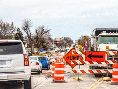 Road construction zone with traffic barrels, warning signs, and a work truck, as vehicles pass through a narrowed lane, illustrating potential hazards and driver-related risks to construction workers.