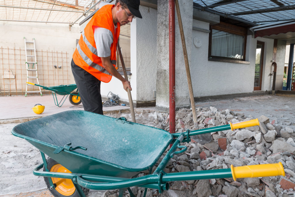 Home repair. Removal of debris, concrete blocks and bricks from the bottom of a roof - terrace. Worker with orange reflective jacket and shovel in his hand