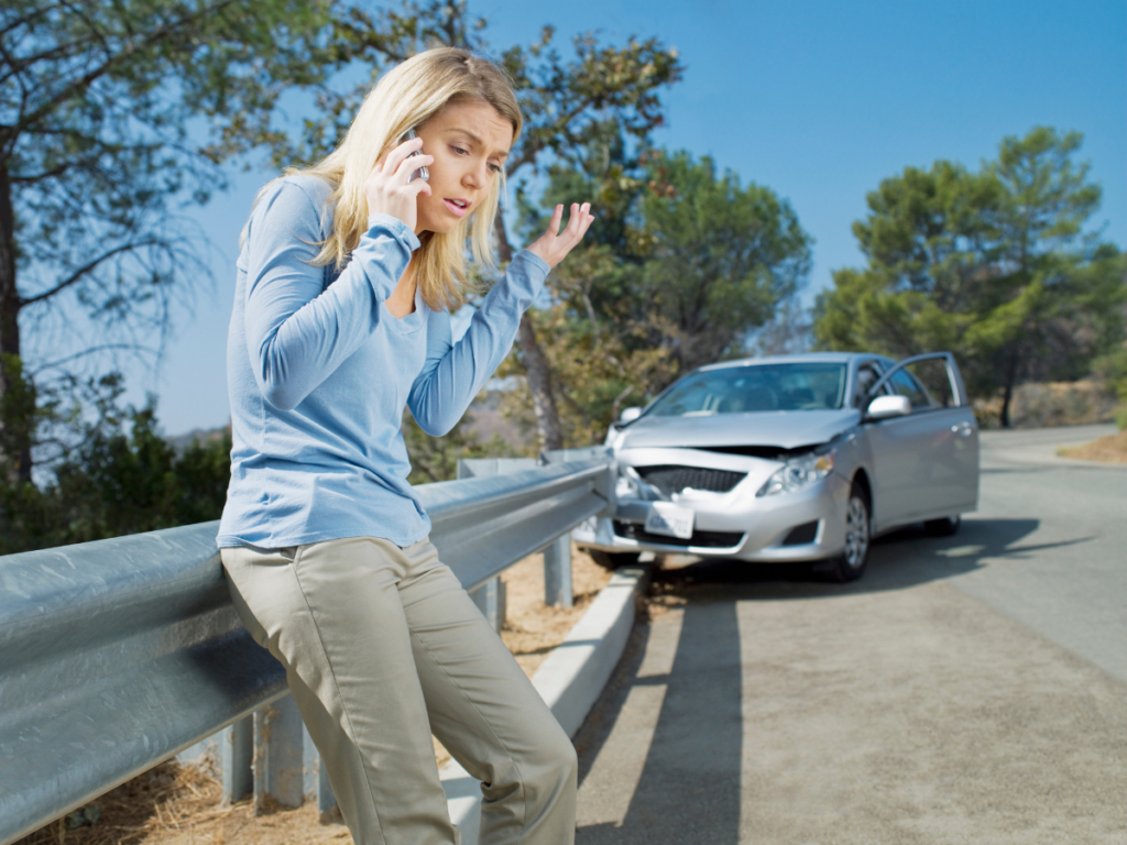 Woman looking stressed on the phone on the side of the road, behind her is her car crashed into the guardrail.