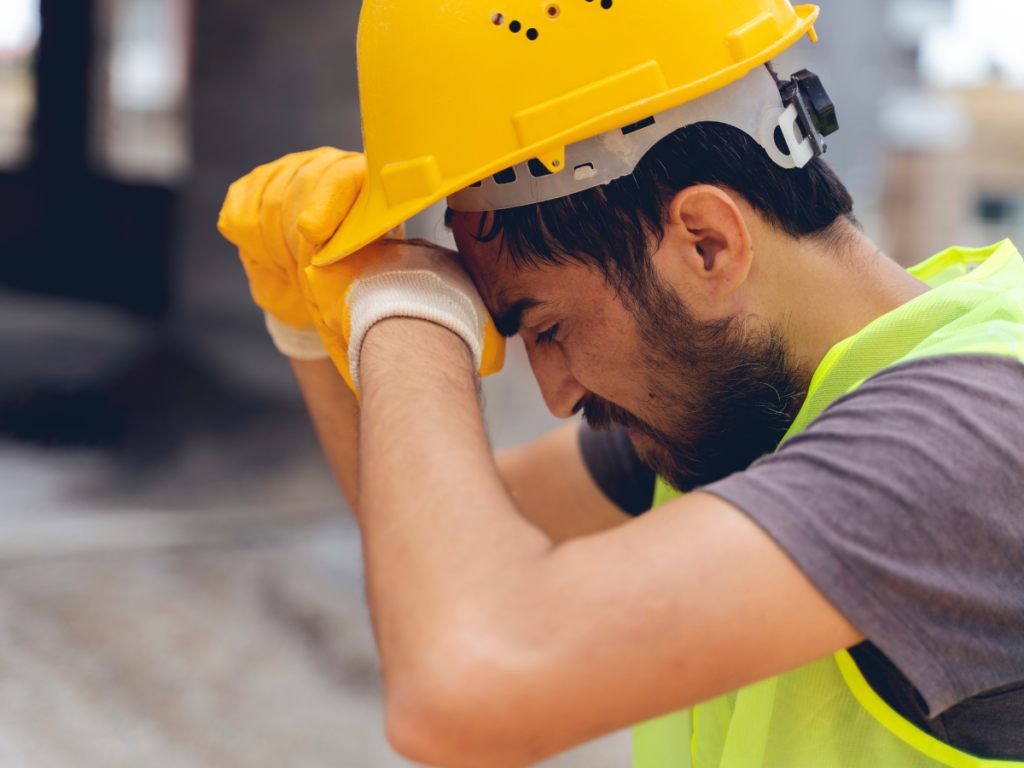 A construction worker in a hard hat with his eyes closed and his hands wiping sweat from his head.