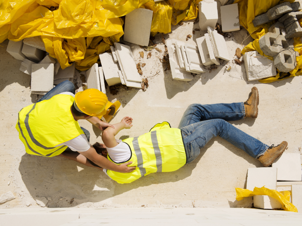 Two construction workers wearing safety vest and hard hats on a construction site. One is on the ground injured and the other is attending to him.